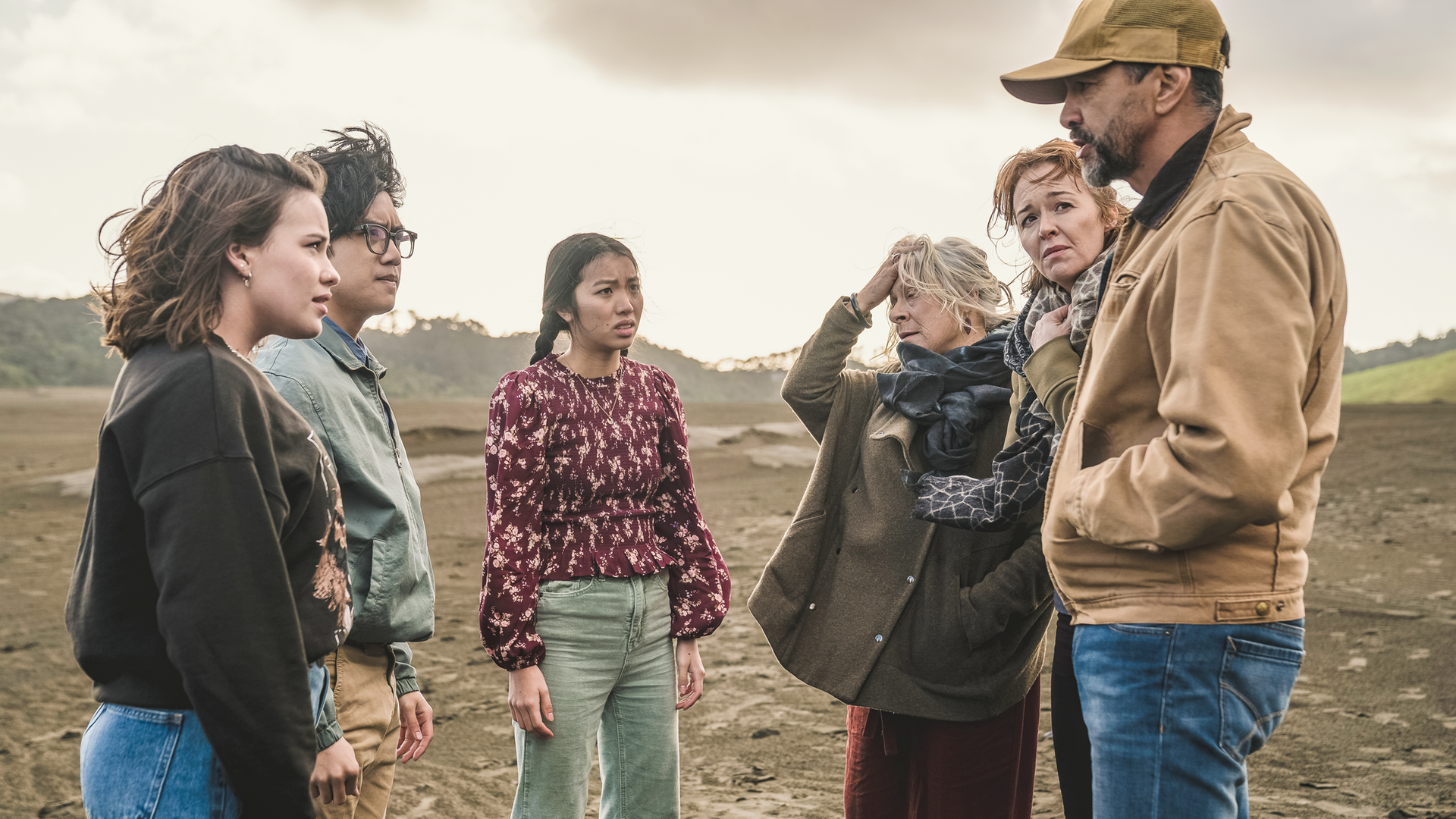 Im sandigen Gelände stehen Issie, Caleb und Caroline gegenüber von Mitch, Amanda und Tom. Alle schauen besorgt. Der Himmel ist grau.