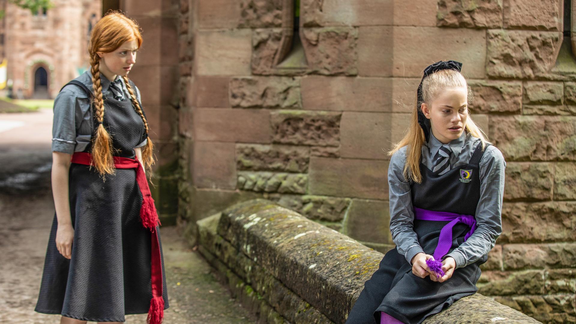 Draußen auf dem Gelände der Graustein-Akademie stellt Mildred, die auf einer Mauer sitzende Esther zur Rede. Esther hat sich seitlich abgewandt und ihre Hände greifen nach ihrer lilafarbenen Schärpe.