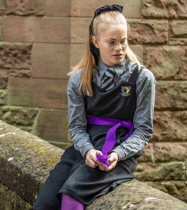 Draußen auf dem Gelände der Graustein-Akademie stellt Mildred, die auf einer Mauer sitzende Esther zur Rede. Esther hat sich seitlich abgewandt und ihre Hände greifen nach ihrer lilafarbenen Schärpe.