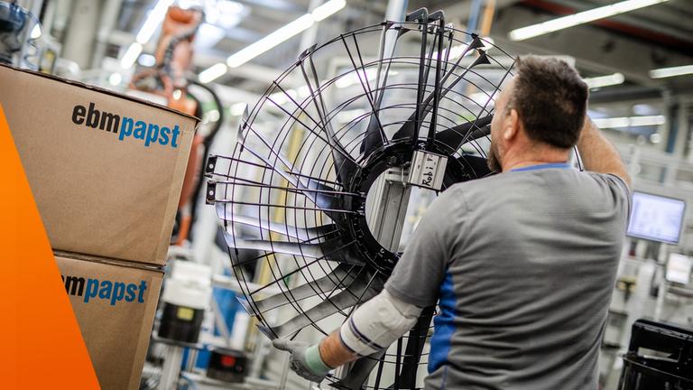 Arbeiter von hinten mit großem Ventilator-Rad in der Hand. Daneben Kartons von EBM-Papst