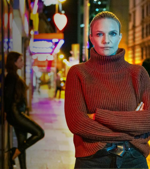 Sarah Tacke steht vor der Kulisse eines Rotlichtviertels. Im Hintergrund lehnt eine Frau in Lederoutfit an einer Wand, ein Mann läuft die Straße entlang.