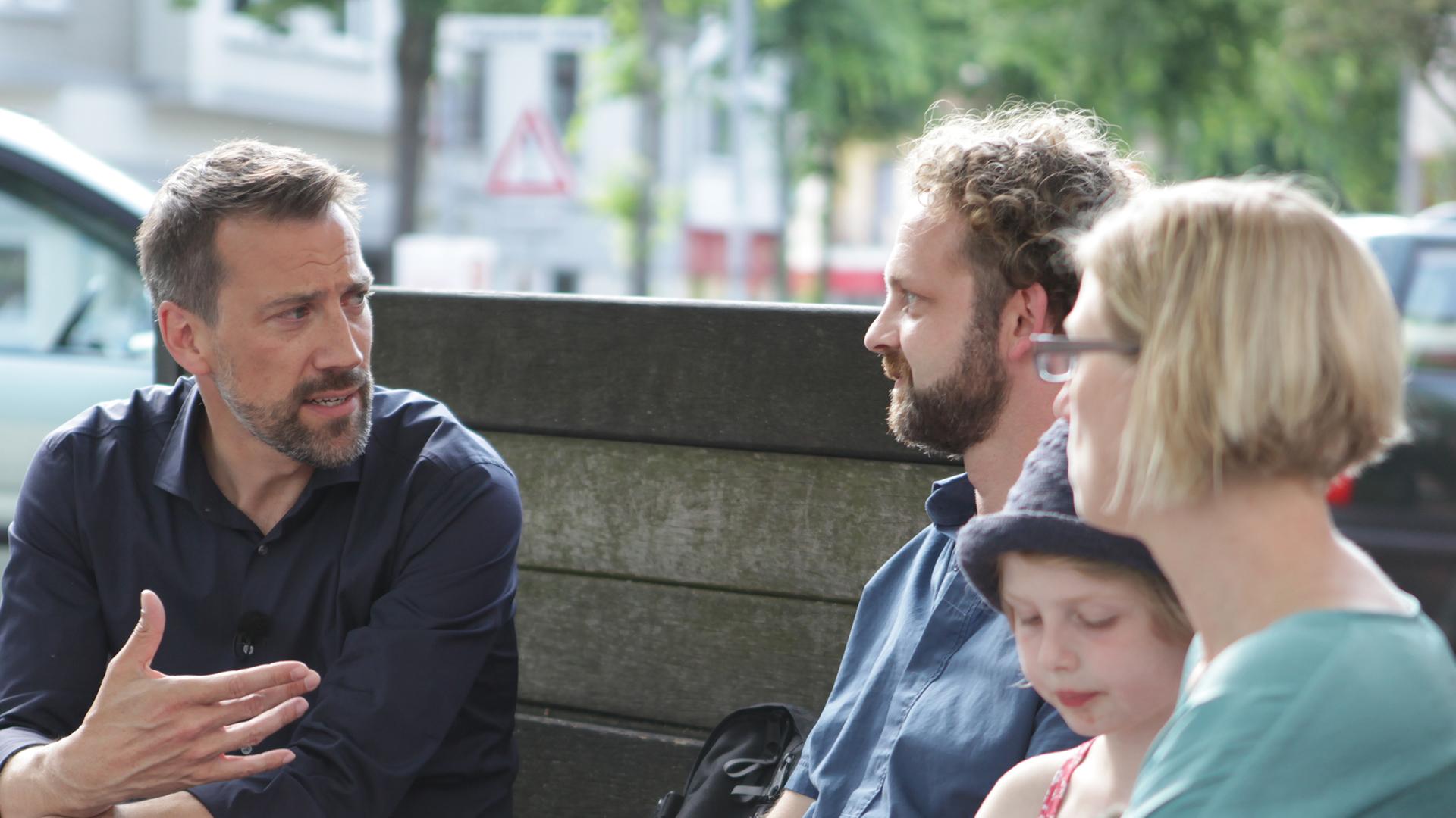 Marcus Niehaves (l) sitzt neben einer Familie auf einer Bank und unterhält sich mit ihr.