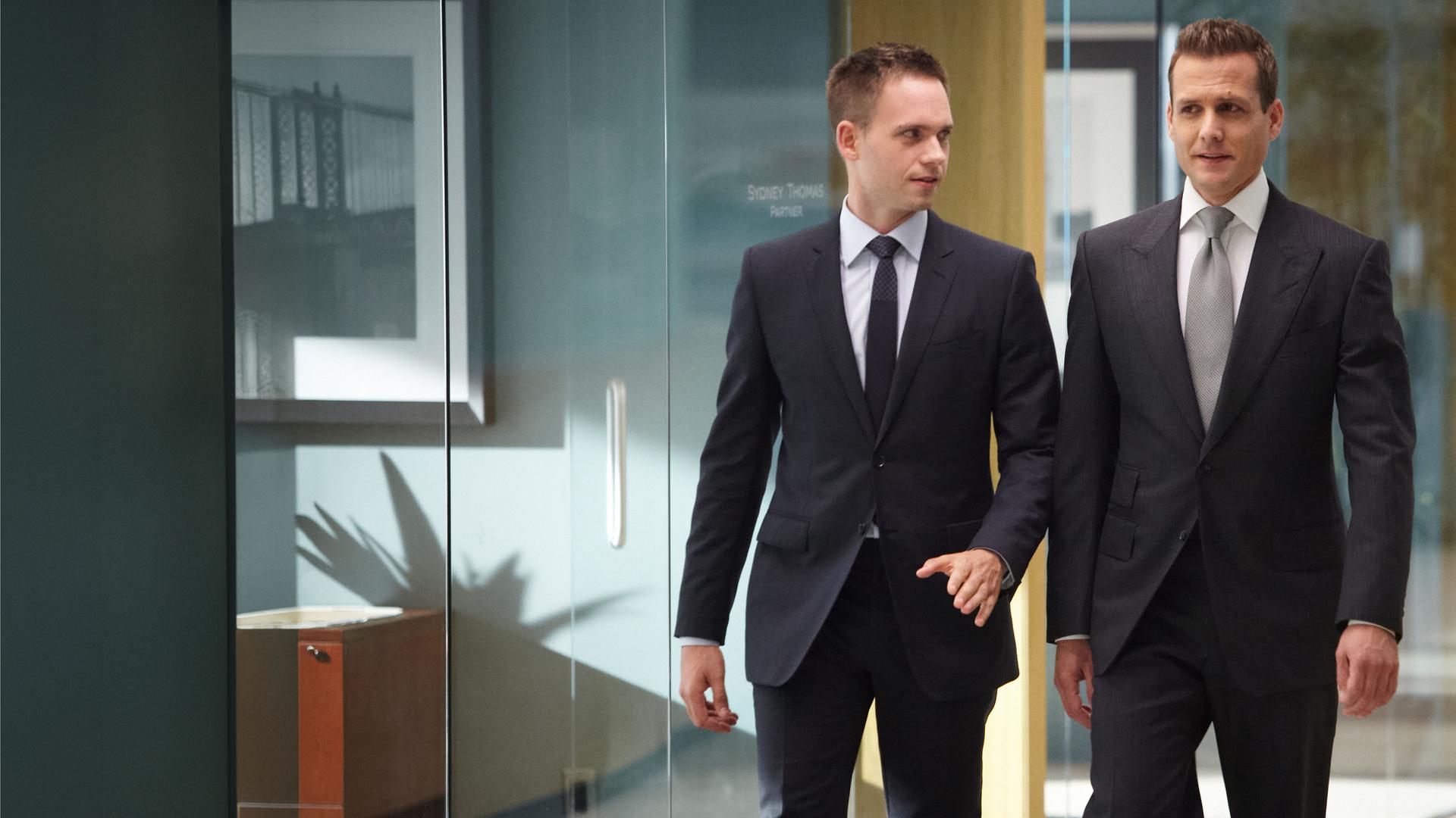 "Suits - Verlieren, um zu gewinnen": Mike (Patrick J. Adams) und Harvey (Gabriel Macht) gehen nebeneinander durch einen gläsernen Büroflur. Beide tragen dunkle Anzüge; Mike schaut zu Harvey, während dieser geradeaus blickt.