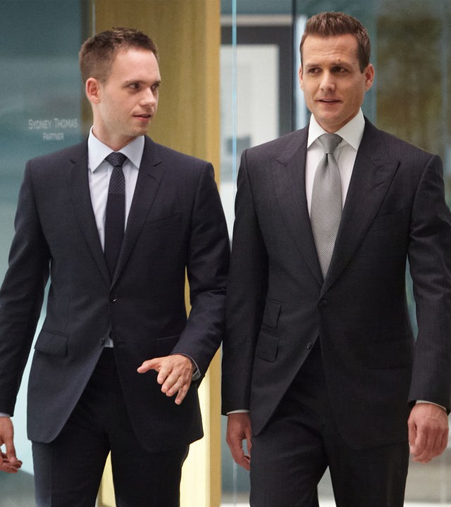 "Suits - Verlieren, um zu gewinnen": Mike (Patrick J. Adams) und Harvey (Gabriel Macht) gehen nebeneinander durch einen gläsernen Büroflur. Beide tragen dunkle Anzüge; Mike schaut zu Harvey, während dieser geradeaus blickt.