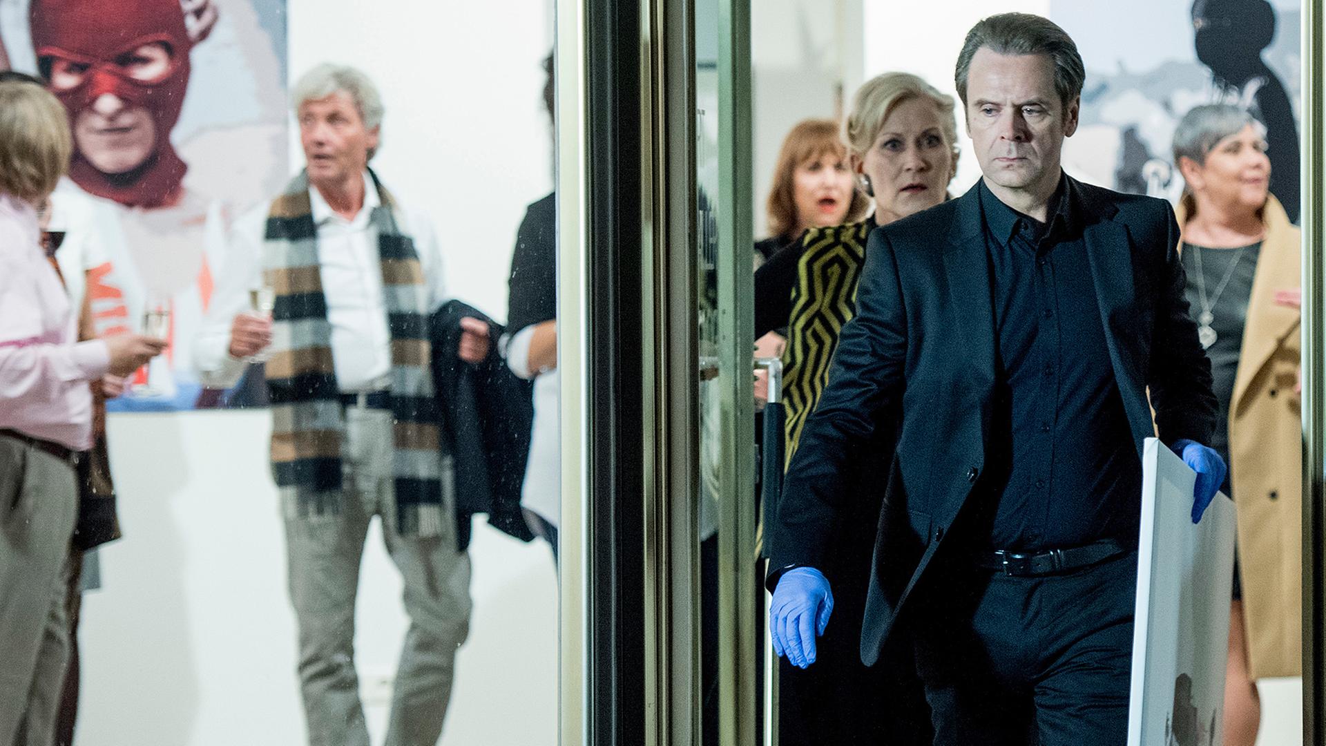 Professor T. (Matthias Matschke, M.) entwendet ein Gemälde, das ihn als Kind zeigt, aus der Galerie seiner Mutter Martha Schönfeld (Hedi Kriegeskotte 4.v.l.).