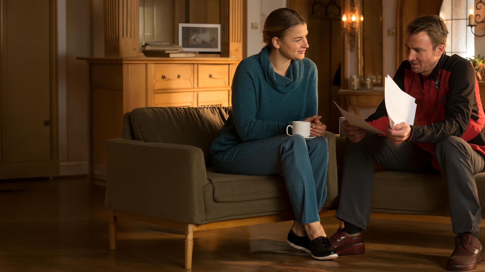 Anne (Ines Lutz) sitzt mit Hans (Heiko Ruprecht) am Tisch und schaut ihn erwartungsvoll an, während er sich einen Stapel Papiere ansieht.