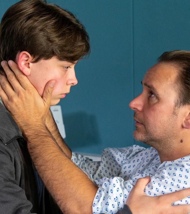 Andreas Wolf (Atef Vogel) sitzt aufrecht in seinem Bett im EKH. Er hat sich zu seinem Sohn Moritz (Jonathan Aaron Finley Ohlrogge) gewandt und hält dessen Kopf liebevoll in seinen Händen. Beide sehen sich traurig in die Augen.