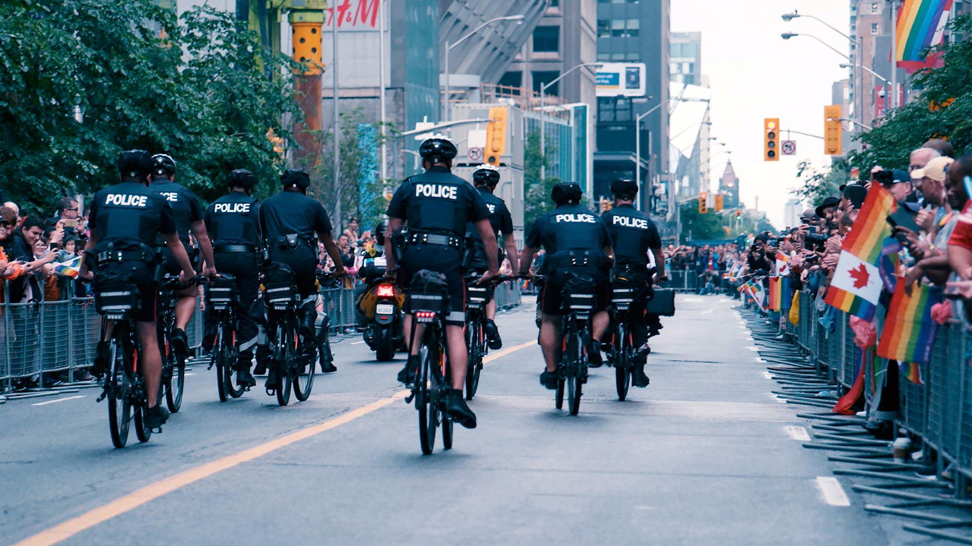 "Vermisst im Gay Village – Serienmorde in Toronto": Acht Polizeibeamte fahren mit Fahrrädern auf einer abgesperrten und von Menschen flankierten Straße. Zuschauer halten Regenbogenflaggen.