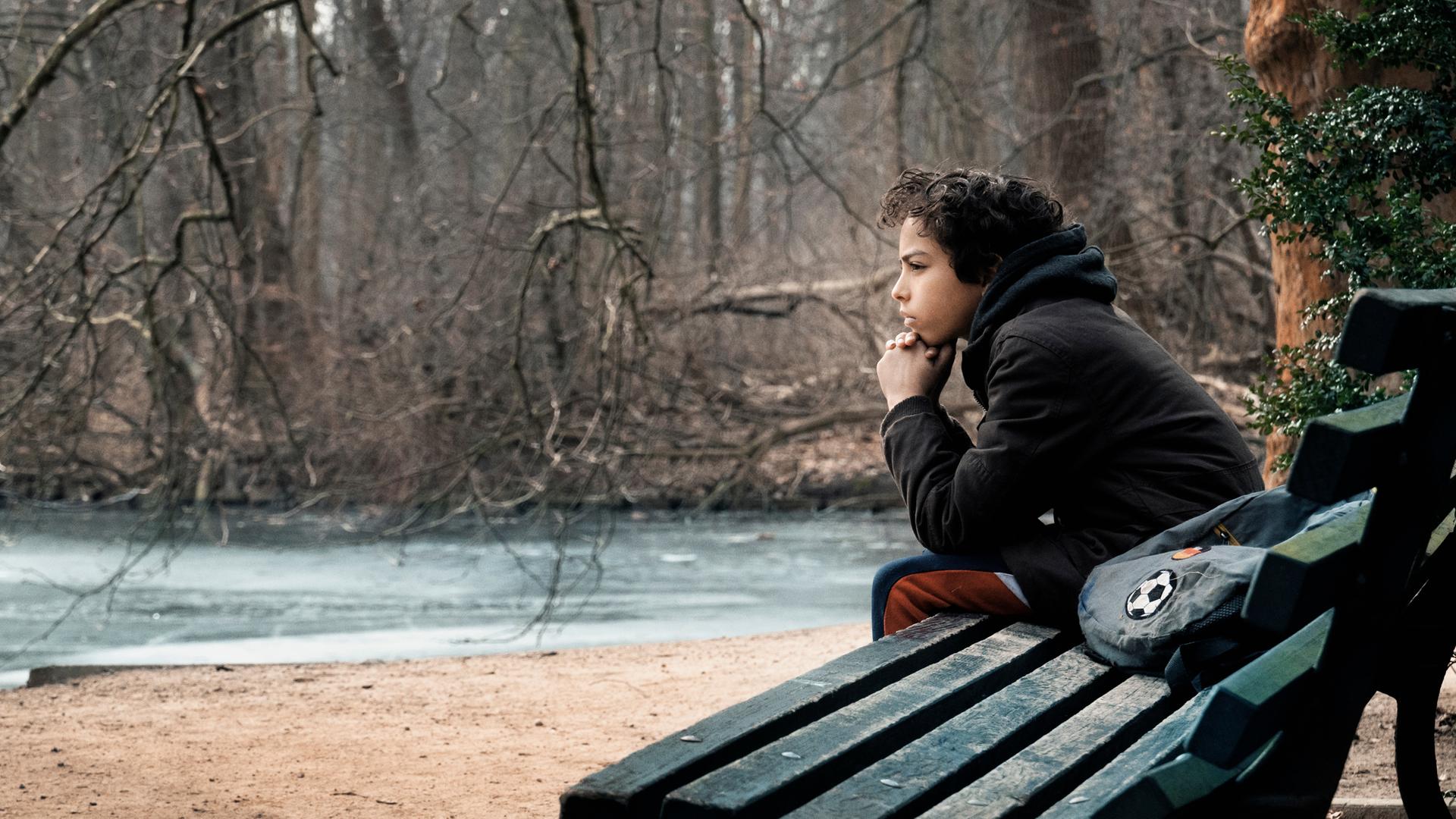 Der kleine Djamal sitzt auf einer Bank in einem winterlichen Park. Er schaut verdrossen auf einen gefrorenen See