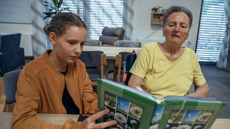 Veronika und ihre Oma sitzen an einem Tisch. Veronika hält ein Buch mit Bildern aus der Natur in der Hand, das sie der Oma zeigt. Im Gesicht der Oma sind deutlich die Spuren der Demenz zu erkennen. Der Raum, den man im Hintergrund wahr nimmt, ist ein mit Sitzmöbeln ausgestatteter Aufenthaltsraum im Altersheim. Alles strahlt Ruhe aus und man erahnt das innige Verhältnis zwischen Oma und Enkelin.
