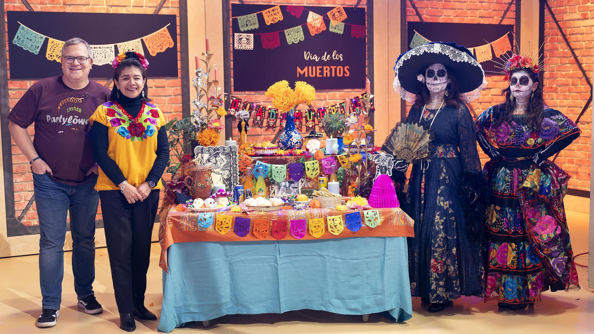 Elton steht zusammen mit Flor Cortés Kargl und zwei geschminkten "Catrinas" im Studio neben einem, unter anderem mit Blumen, Fotos und Kerzen, bunt geschmückten Altar.