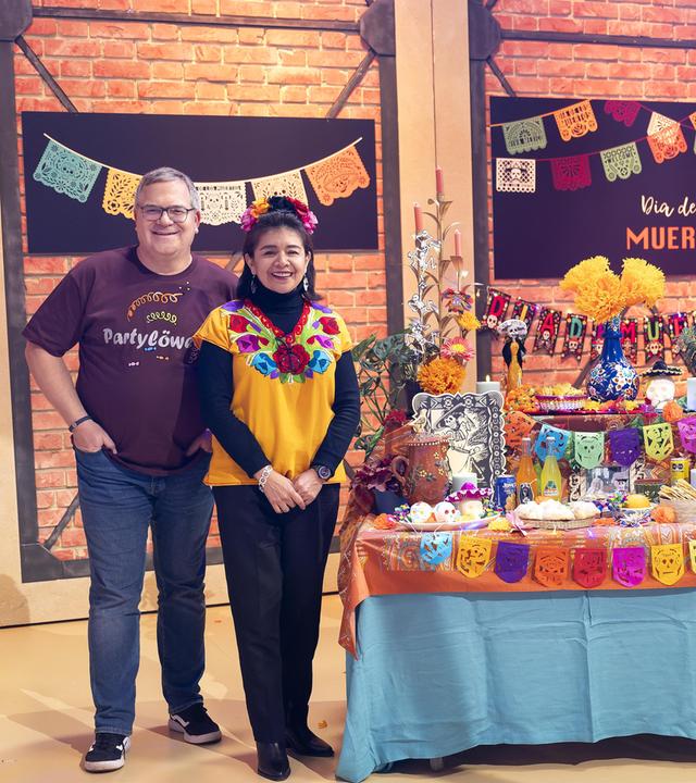 Elton steht zusammen mit Flor Cortés Kargl und zwei geschminkten "Catrinas" im Studio neben einem, unter anderem mit Blumen, Fotos und Kerzen, bunt geschmückten Altar.