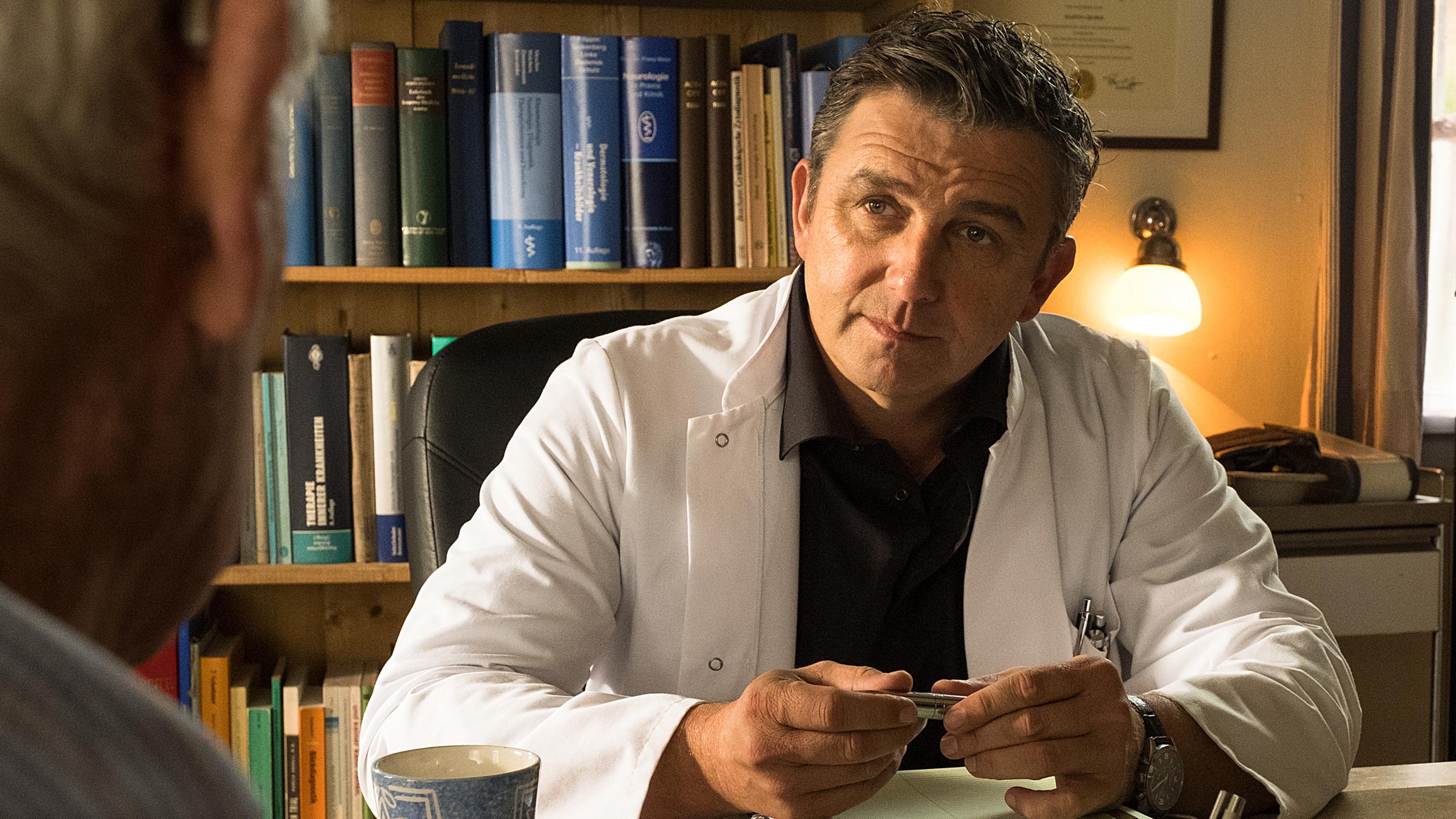 In der Arztpraxis sitzt Dr. Martin Gruber (Hans Sigl) an seinem Schreibtisch und unterhält sich mit Roman Melchinger (Siegfried Rauch), der ihm gegenübersitzt.