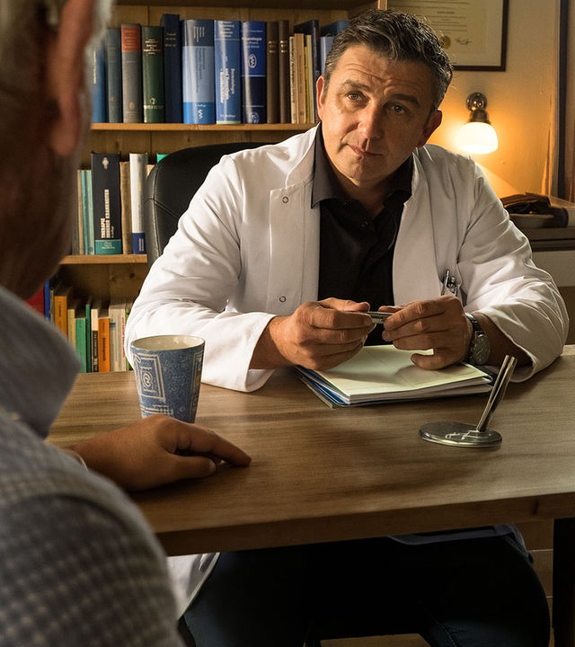 In der Arztpraxis sitzt Dr. Martin Gruber (Hans Sigl) an seinem Schreibtisch und unterhält sich mit Roman Melchinger (Siegfried Rauch), der ihm gegenübersitzt.