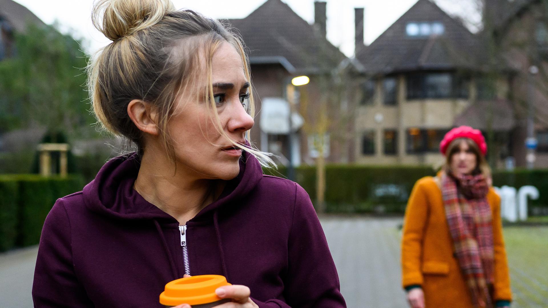 Betty (Annina Hellenthal) steht mit einem Kaffeebecher auf dem Parkplatz vor der Klinik. Sie schaut verängstigt und angespannt über ihre Schulter. Rike Köhler (Isabell Horn) steht hinter ihr, ihr Fahrrad liegt auf dem Boden.