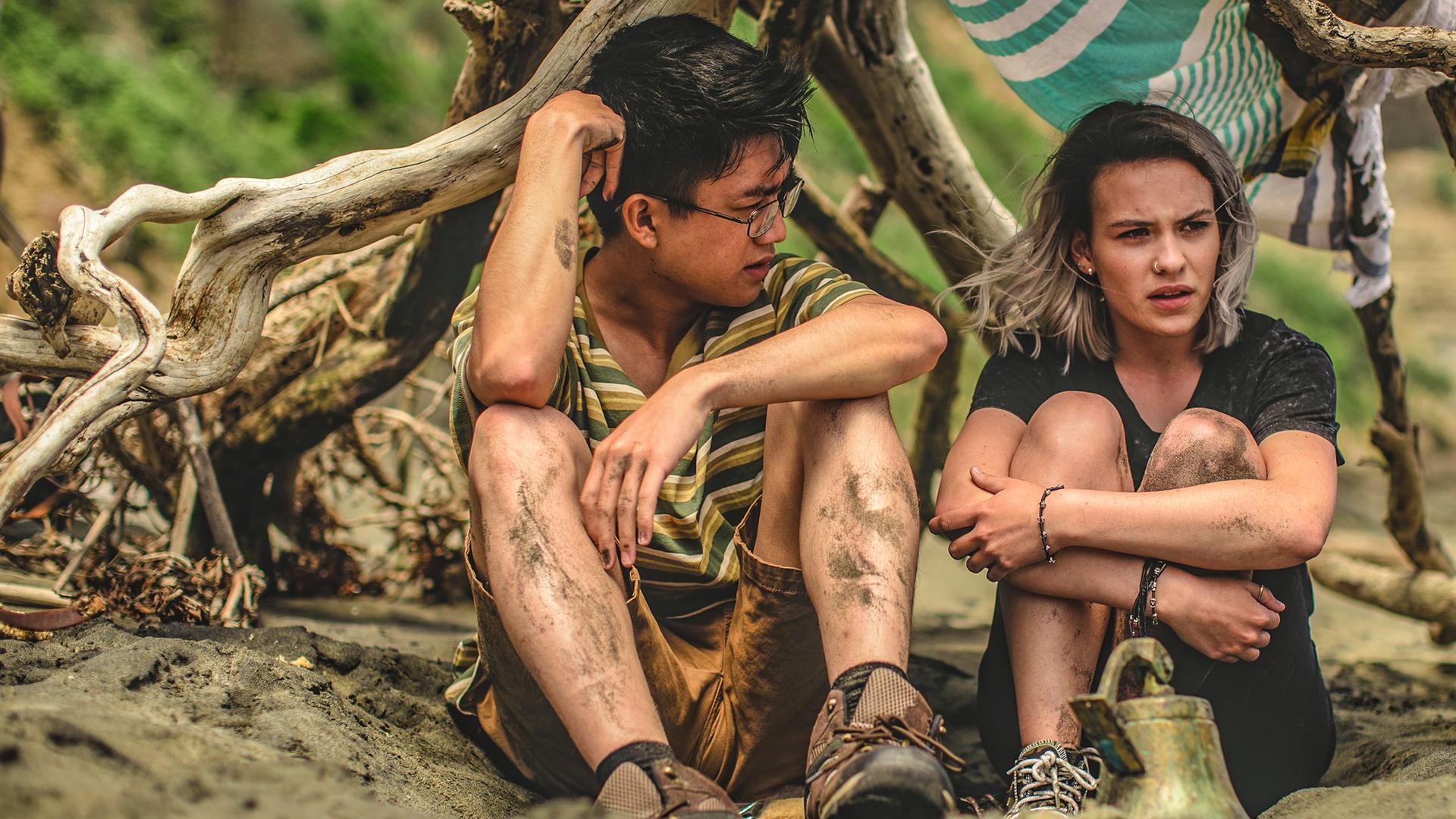 Issie und Caleb sitzen gemeinsam in einem selbst gebauten Zelt am Strand. Beide haben die Knie zum Oberkörper gezogen und sehen nachdenklich aus.