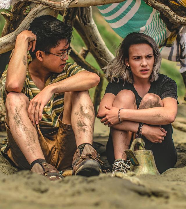 Issie und Caleb sitzen gemeinsam in einem selbst gebauten Zelt am Strand. Beide haben die Knie zum Oberkörper gezogen und sehen nachdenklich aus.