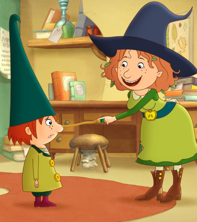 Petronella tippt lachend den Wichtel Nisse mit ihrem Zauberstab an, im Hintergrund liegt ihr aufgeschlagenes Zauberbuch auf einem Ständer in ihrem Zimmer.
