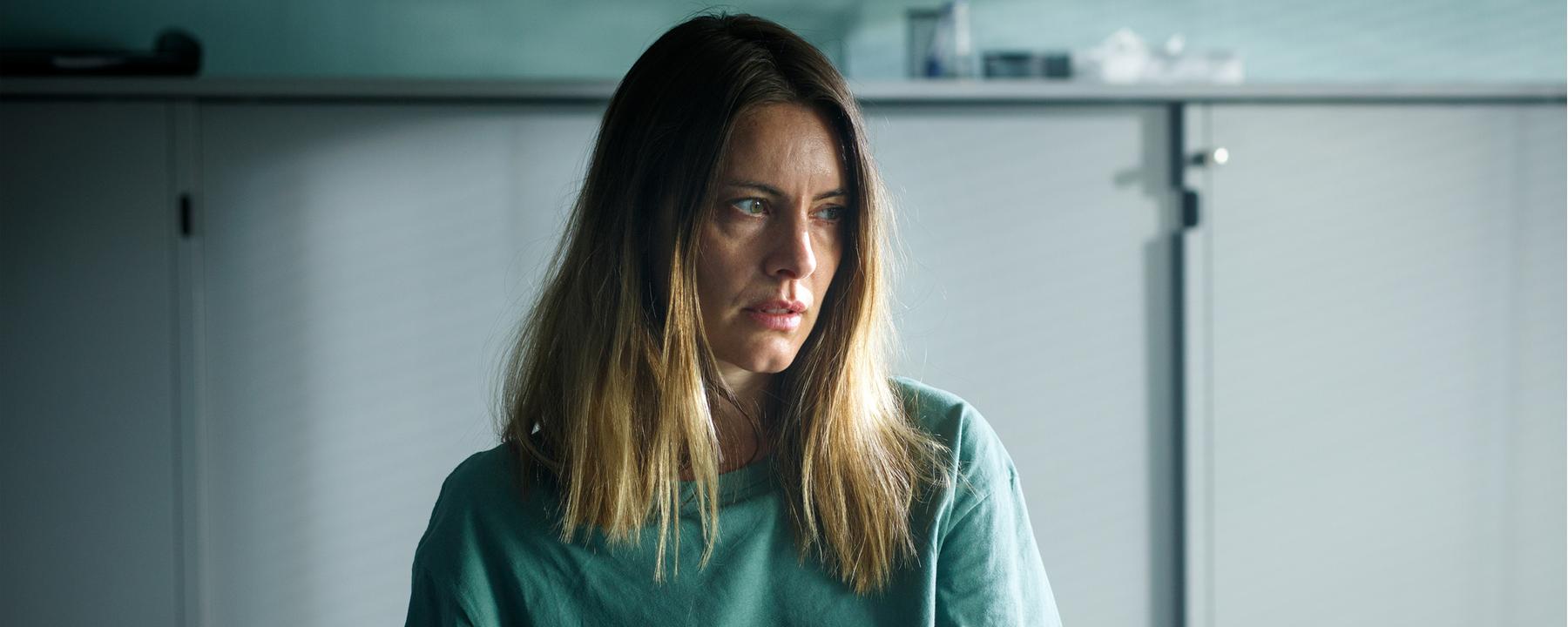 Vicky (Malin Crépin) sitzt an einem Tisch und sieht nachdenklich zur Seite. Im Hintergrund steht ein grauer Aktenschrank.