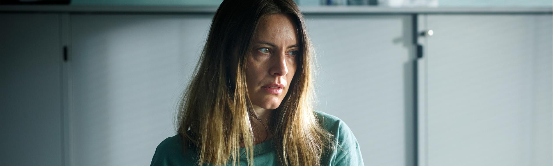 Vicky (Malin Crépin) sitzt an einem Tisch und sieht nachdenklich zur Seite. Im Hintergrund steht ein grauer Aktenschrank.
