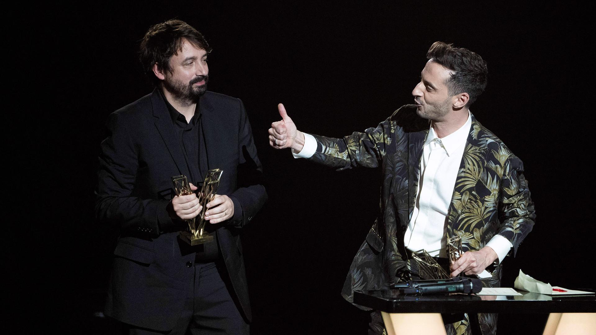 Ein Mann in dunklem Anzug steht auf einer Bühne und hält einen Preis in der Hand. Rechts neben ihm steht ein Mann an einem Pult.