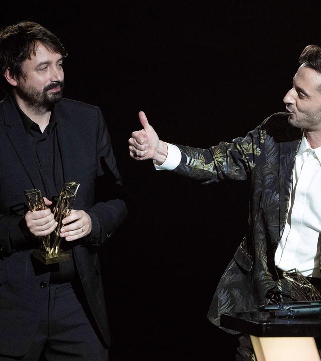 Ein Mann in dunklem Anzug steht auf einer Bühne und hält einen Preis in der Hand. Rechts neben ihm steht ein Mann an einem Pult.