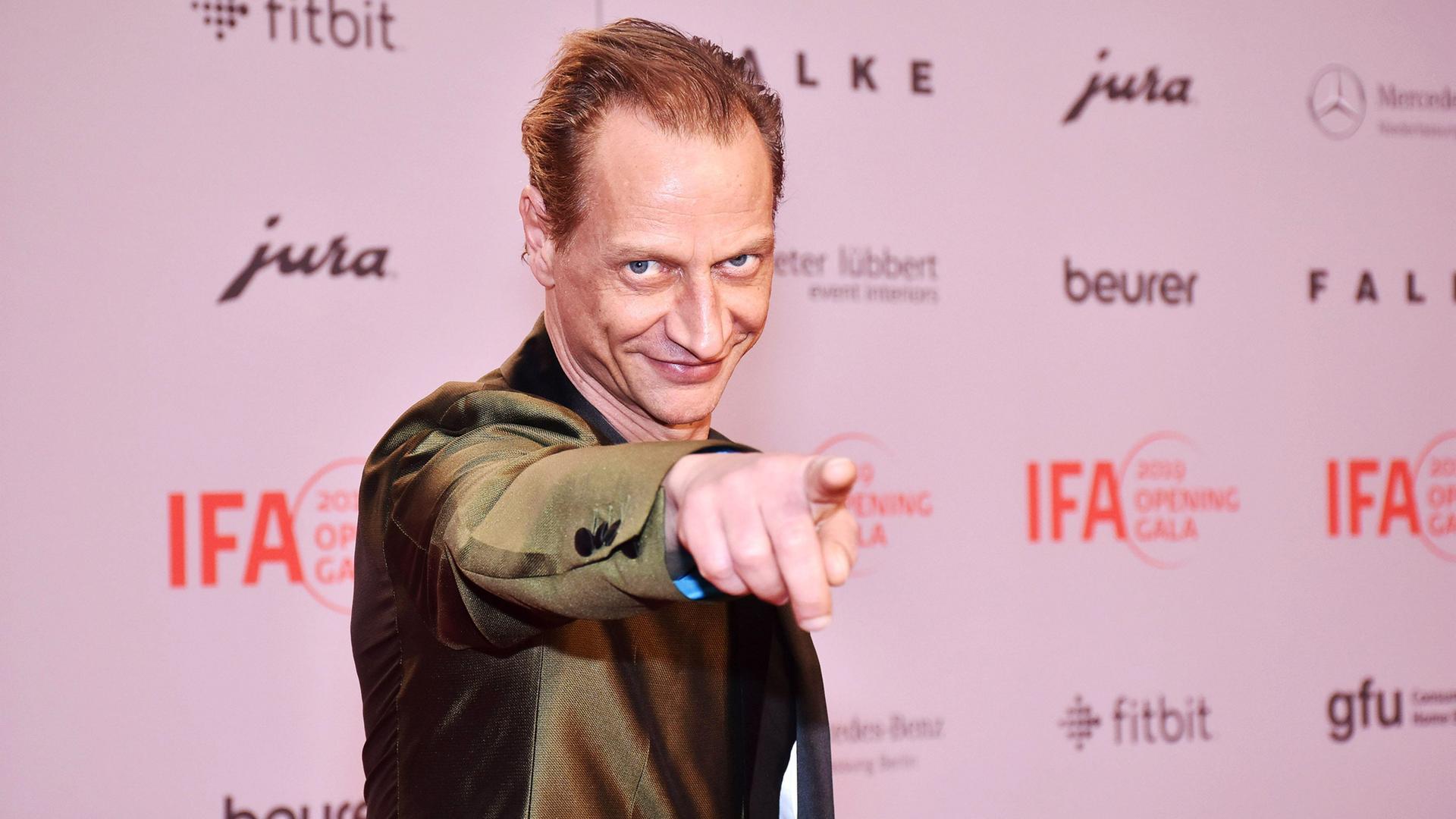 Foto von einem Mann im Anzug, der verschmitzt lächelnd mit dem Finger auf den Betrachter zeigt.