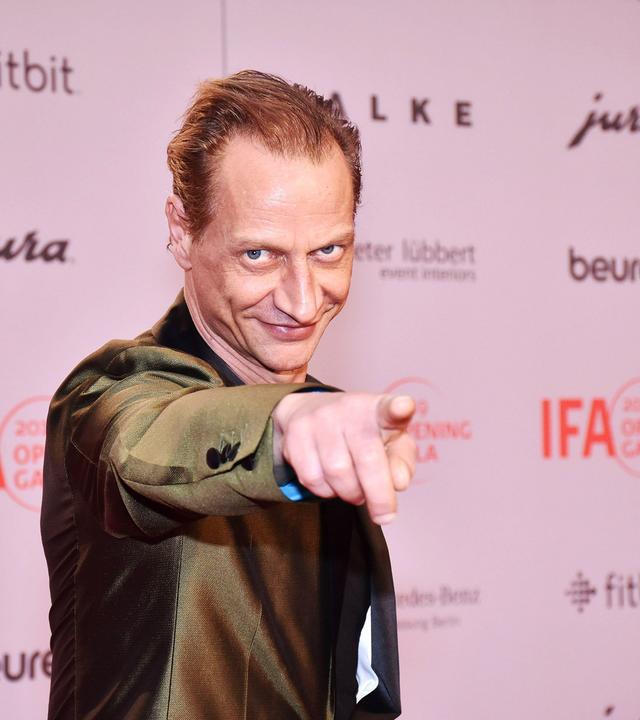 Foto von einem Mann im Anzug, der verschmitzt lächelnd mit dem Finger auf den Betrachter zeigt.