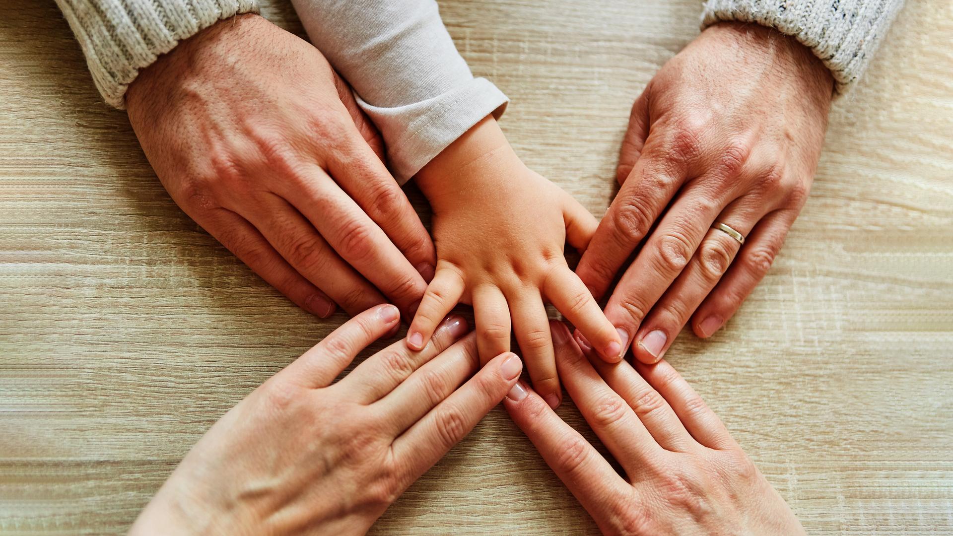 Fünf Hände kommen von unterschiedlichen Seiten ins Bild und berühren sich in der Mitte. Die Hände eines Mannes, einer Frau und eine Kinderhand.