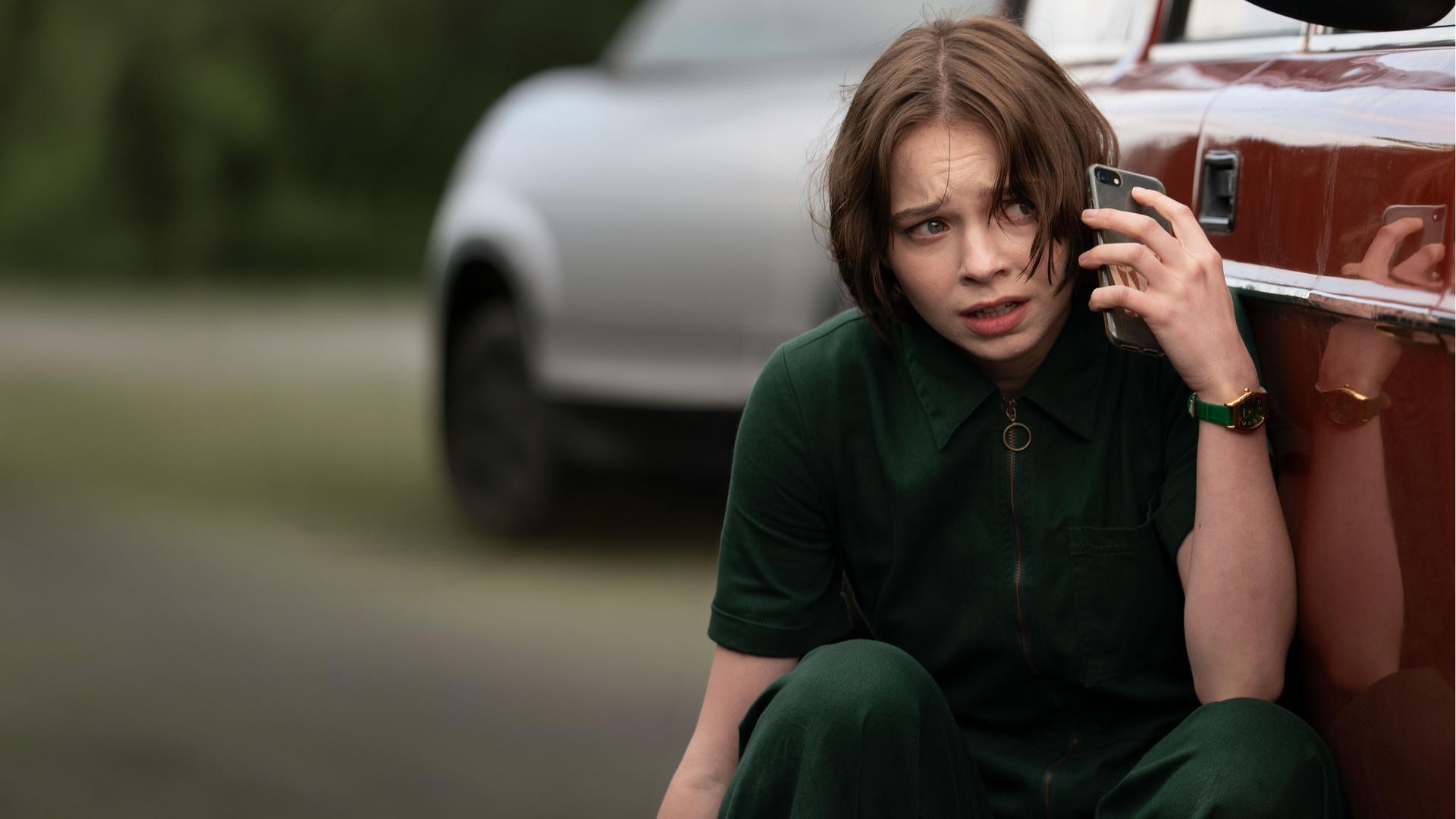 "A Good Girl’s Guide to Murder – Vielleicht kann man gar nicht gut sein": Pip Fitz-Amobi (Emma Myers) hockt dicht neben ihrem roten Auto und hält ein Handy ans Ohr. Ihr Blick ist besorgt.