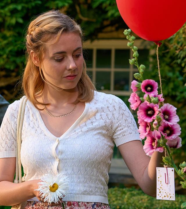 Joey (Bernd-Christian Althoff) steht mit Gwen (Meriel Hinsching) im Garten. Gwen hält einen roten Luftballon mit einer angebrachten Nachricht in der Hand.