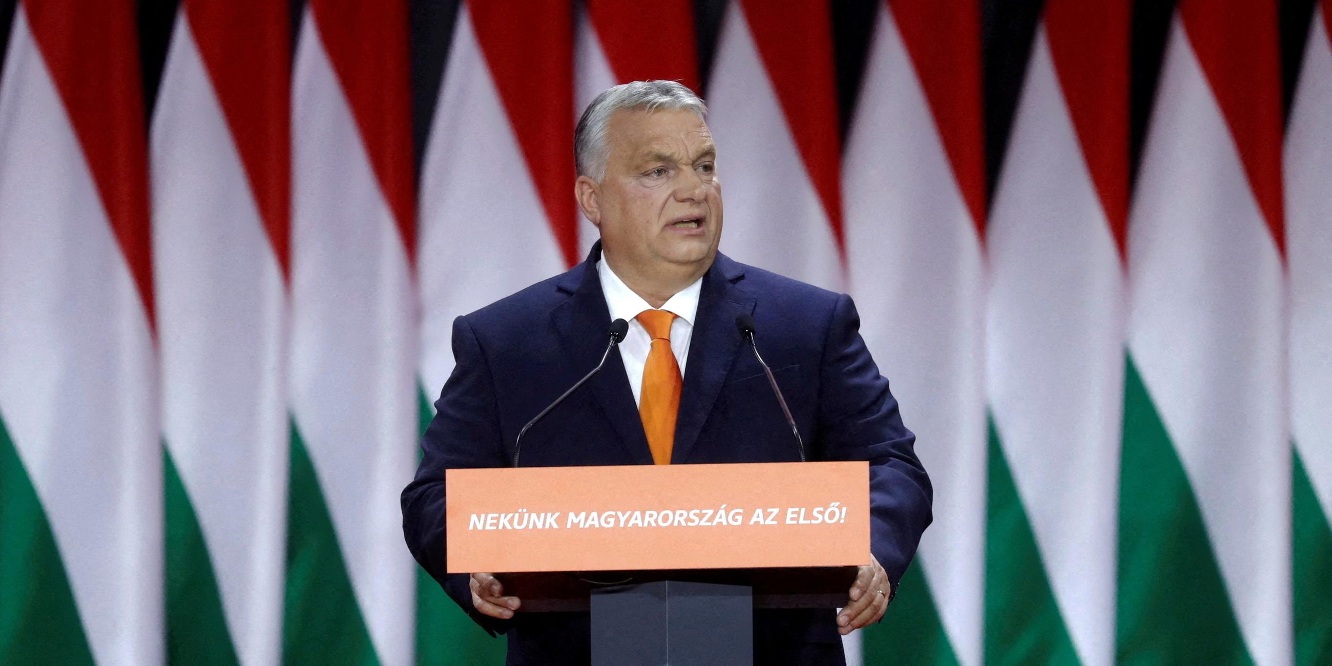Viktor Orban steht vor Ungarn-Flaggen an einem Rednerpult und hält sich mit beiden Händen daran fest.