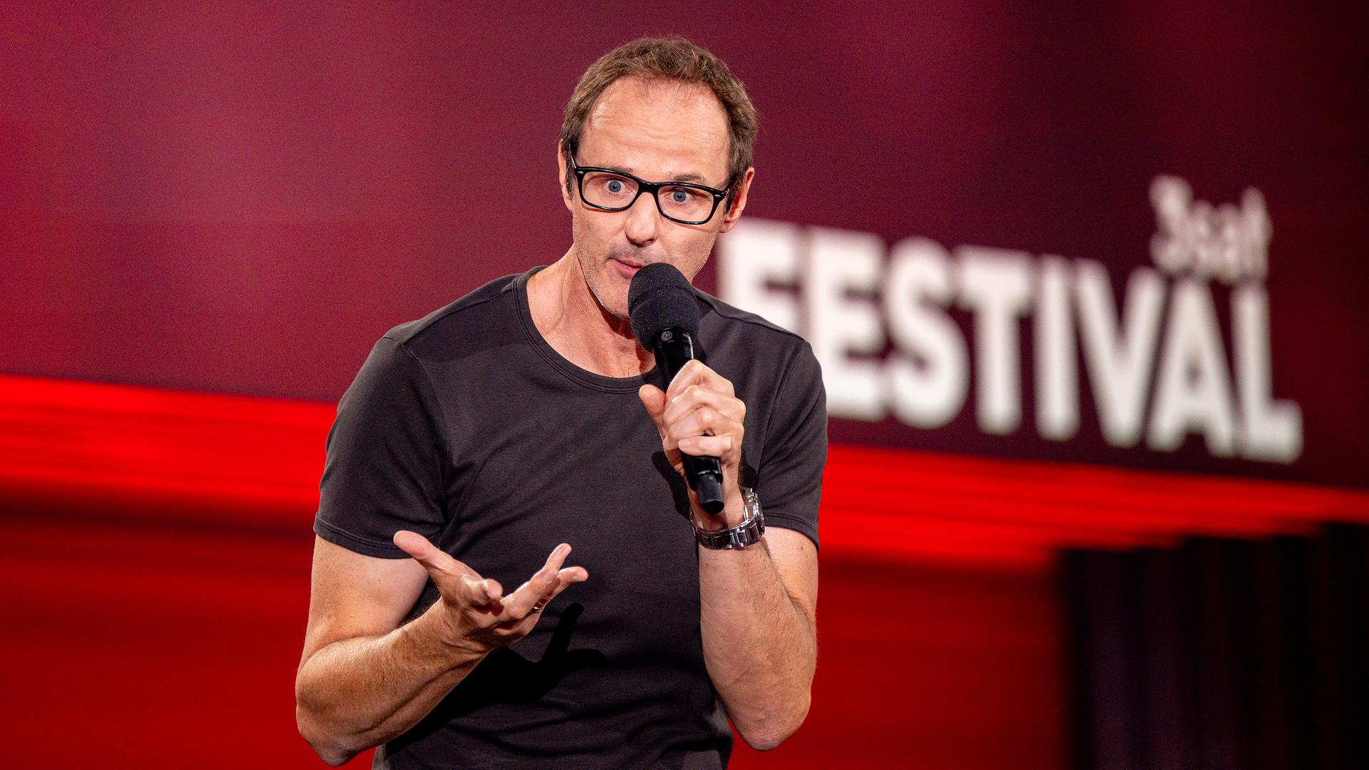 ince Ebert steht in der Mitte des Bildes. Er hebt seine rechte Hand. In der linken Hand hält er sein Mikrofon. Der Hintergrund ist rot beleuchtet.