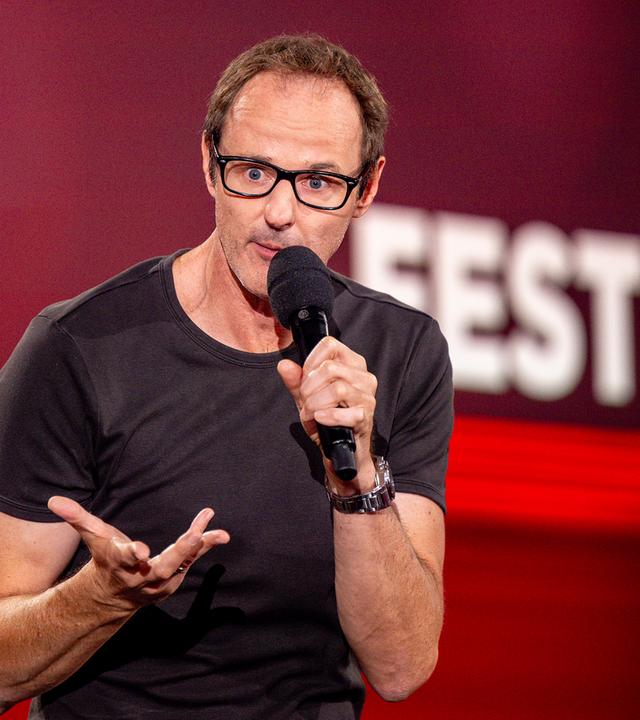 ince Ebert steht in der Mitte des Bildes. Er hebt seine rechte Hand. In der linken Hand hält er sein Mikrofon. Der Hintergrund ist rot beleuchtet.