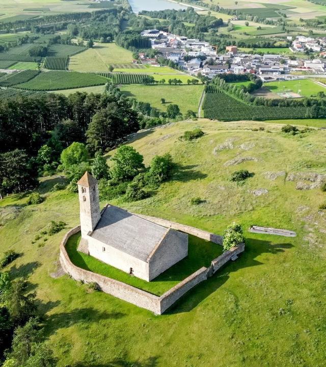 Das Bild zeigt die kleine Kirche Tartscher Buehel, die auf einem sanften Hügel steht. Sie hat eine rechteckige Form mit einem schlichten grauen Dach und einem Glockenturm, der an einer Ecke erhöht ist. Um die Kirche herum befindet sich eine halbkreisförmige Steinmauer. Der Hügel ist von saftigem Grün umgeben, und es gibt einige verstreute Bäume. Im Hintergrund erstreckt sich eine weite Landschaft mit Feldern und Wäldern, die in verschiedenen Grüntönen leuchten. In der Ferne sind einige Gebäude und Straßen zu erkennen, die Teil einer Ortschaft sind. Das Bild vermittelt einen ruhigen, natürlichen Eindruck und zeigt die idyllische alpine Umgebung des Vinschgaus in Südtirol.