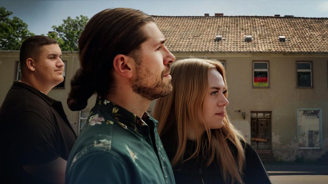 Drei Personen vor einem Wohnhaus in einer Kleinstadt. Sie stehen nebeneinander auf der Straße, während sich das Gebäude über die gesamte Bildbreite im Hintergrund erstreckt.