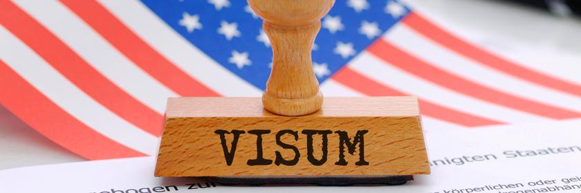 Nahaufnahme eines Visumstempels auf einem Antragsformular. Dahinter sind unscharf die amerikanische Flagge und ein Stift zu sehen. Die Aufnahme steht sinnbildlich für geplante Änderungen der US-Visabestimmungen