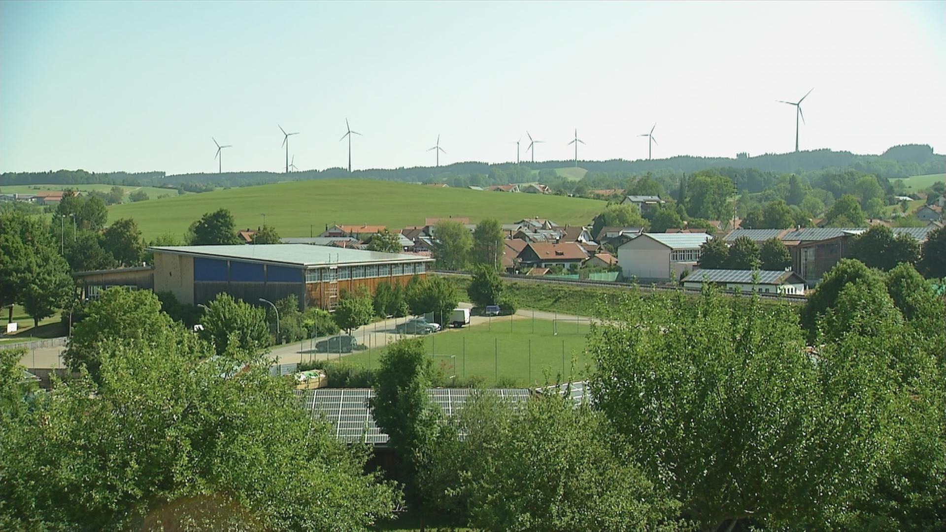 Gebäude mit Solarplatten auf dem Dach und Grünflächen, am Horizont sind Windräder
