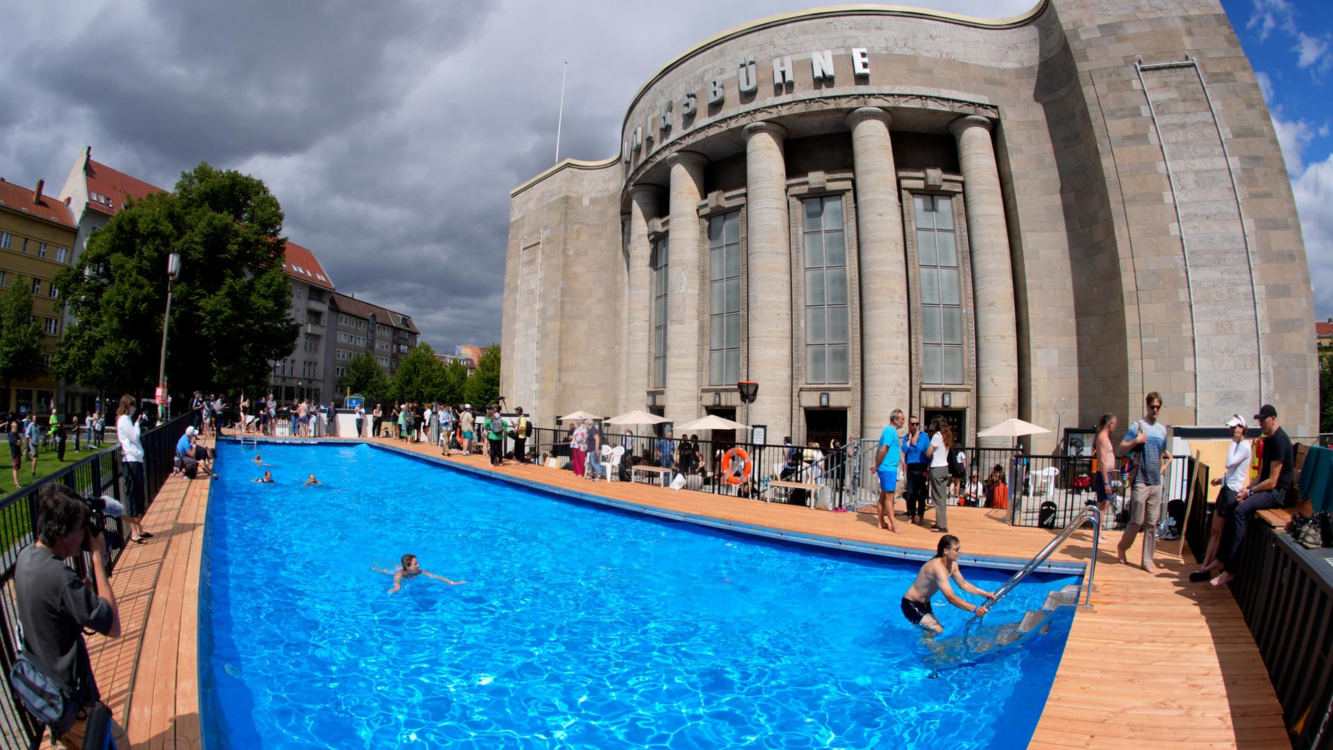 Menschen schwimmen in und stehen an einem Schwimmbad vor einem Theaterbau.