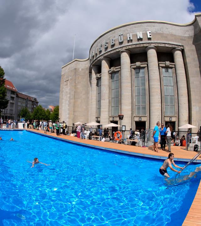 Menschen schwimmen in und stehen an einem Schwimmbad vor einem Theaterbau.