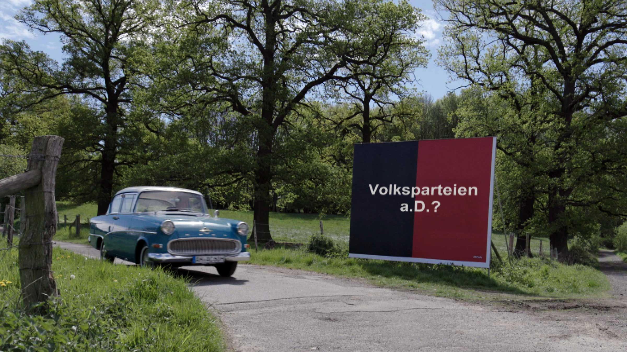 "Volksparteien a.D.? - Zeitenwende in Deutschland": Trabbi auf dem Land unterwegs. Er fährt an einem Plakat vorbei mit der Aufschrift "Volksparteien a. D.?"