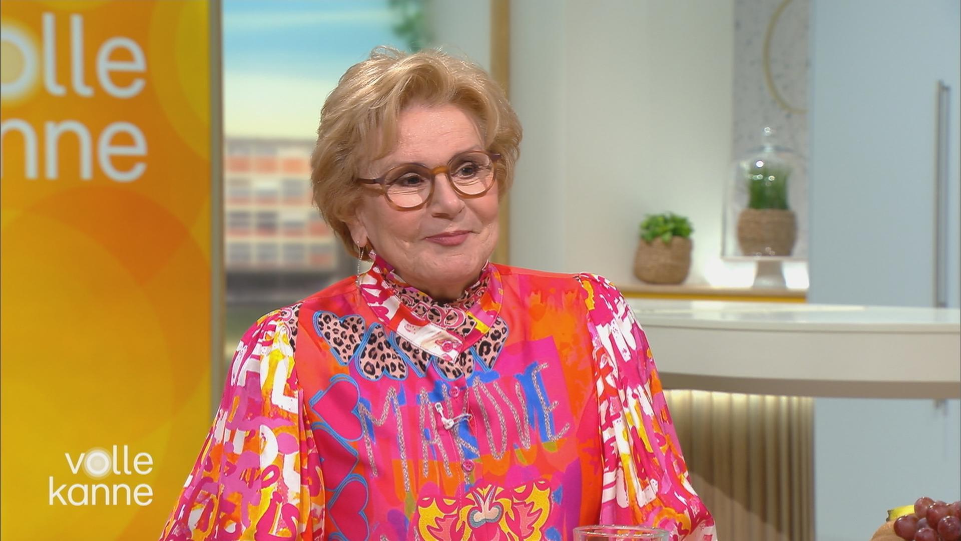 Peggy March sitzt im Volle Kanne Set und trägt ein schrilles Oberteil mit Pink-, Gelb- und Orangetönen und blauen Akzenten. 