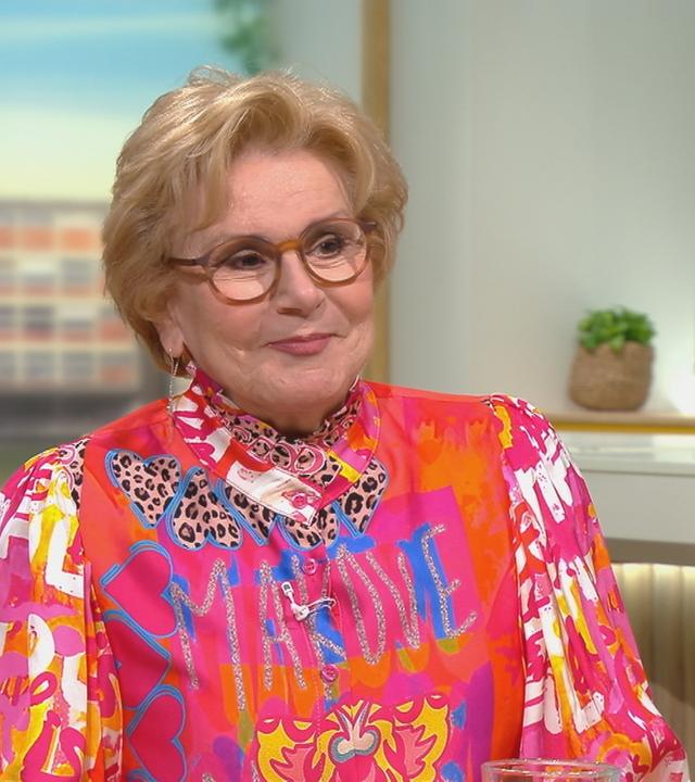 Peggy March sitzt im Volle Kanne Set und trägt ein schrilles Oberteil mit Pink-, Gelb- und Orangetönen und blauen Akzenten. 