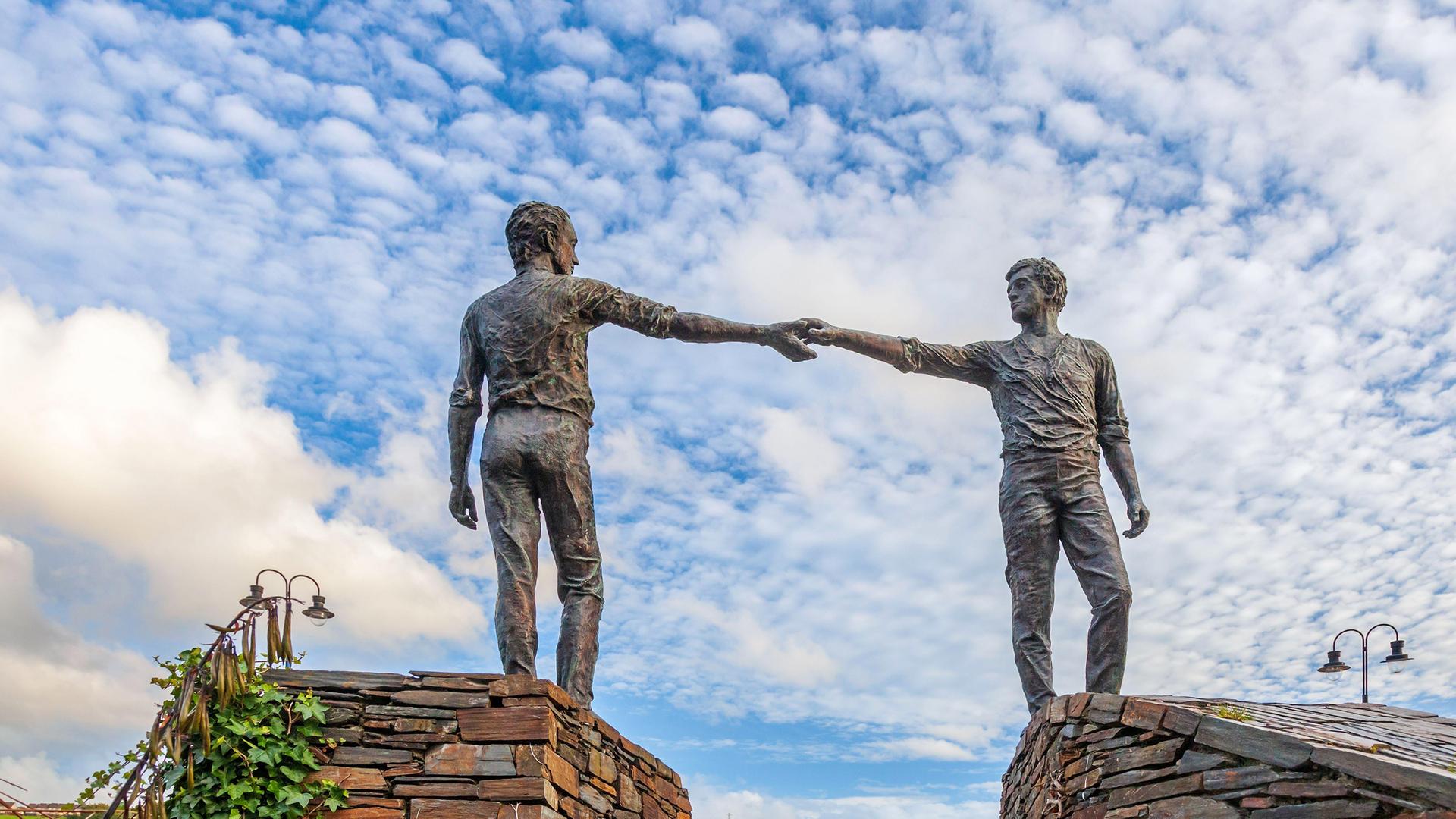 Denkmal zweier Männer, die auf Mauern stehend, sich über den dazwischenliegenden Graben hinweg die Hände reichen.