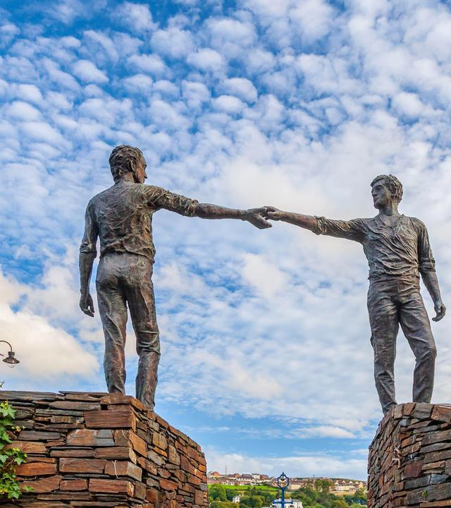 Denkmal zweier Männer, die auf Mauern stehend, sich über den dazwischenliegenden Graben hinweg die Hände reichen.