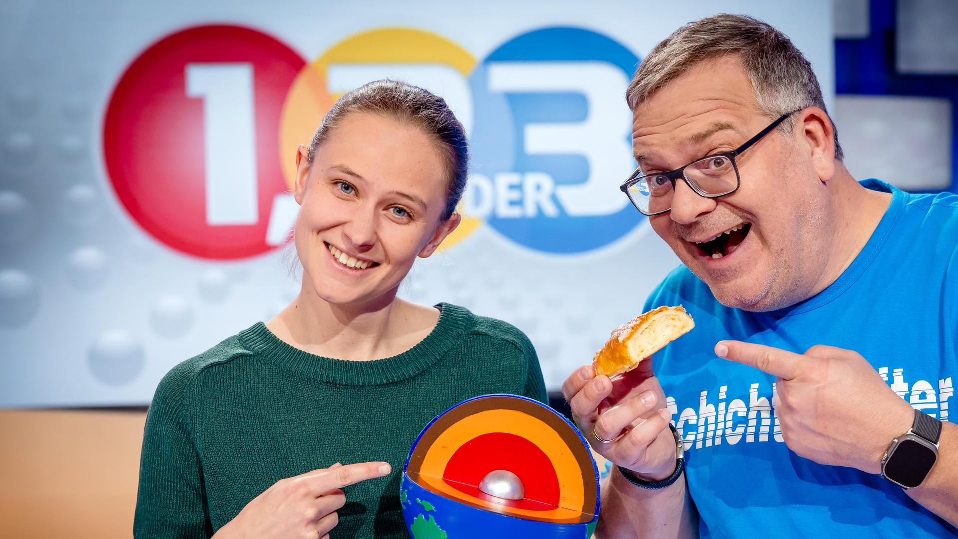 Elton steht mit Geologin Alicia Rohnacher zusammen. Sie hält ein Modell des Erdmantels vor sich. Elton und sie deuten darauf.