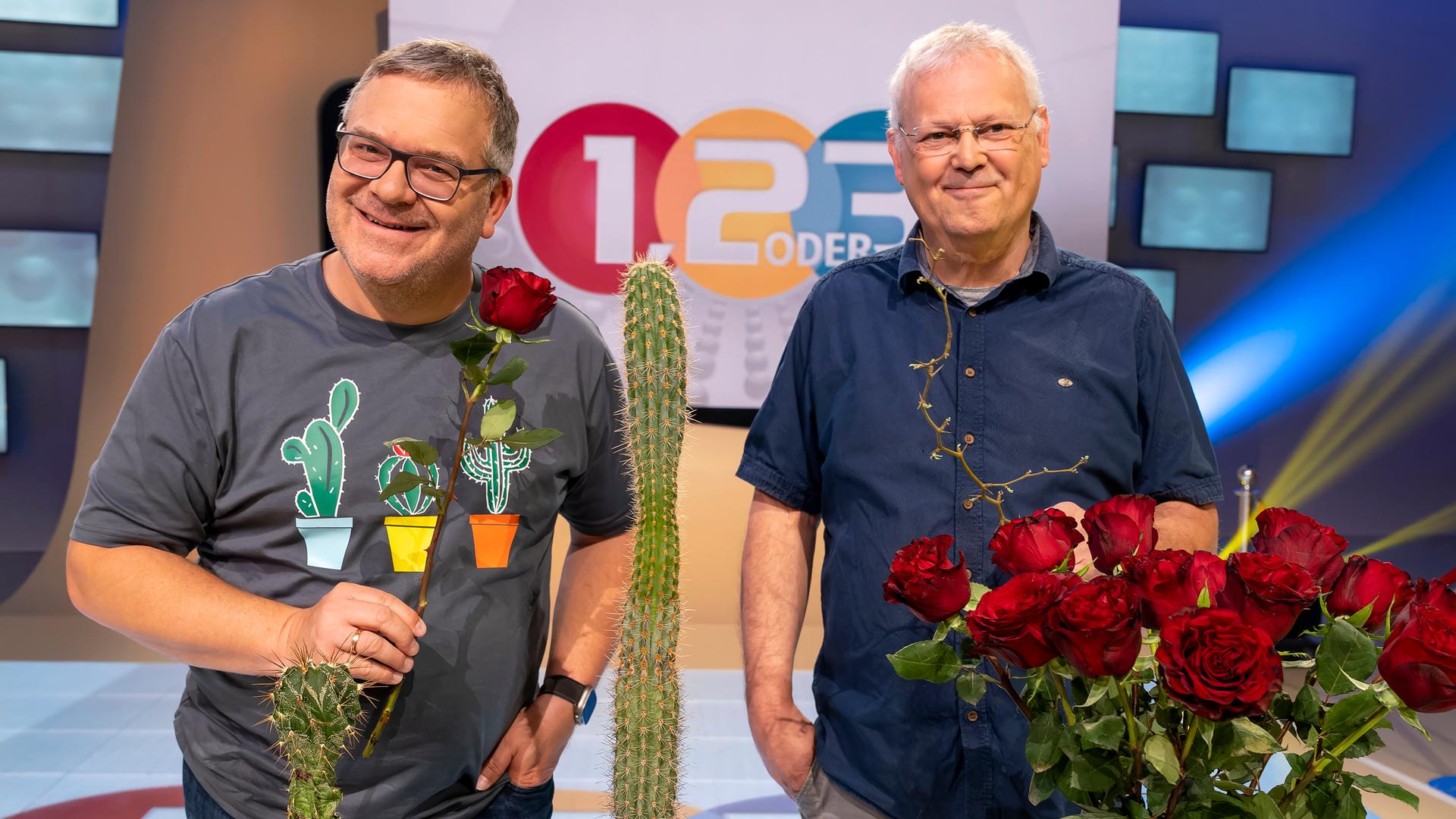 Elton und Professor Christoph Neinhuis stehen hinter einem Tisch, auf dem ein Rosenstrauß in einer Vase und mehrere unterschiedlich große Kakteen platziert sind.