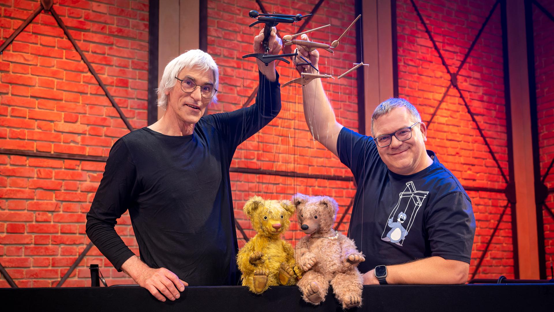 Puppenspieler Detlef Schmelz und Elton stehen nebeneinander und halten Marionetten in den Händen. Die Marionettenpuppen sind zwei Teddybären aus Stoff, die auf einer Kante vor Detlef Schmelz und Elton sitzen.