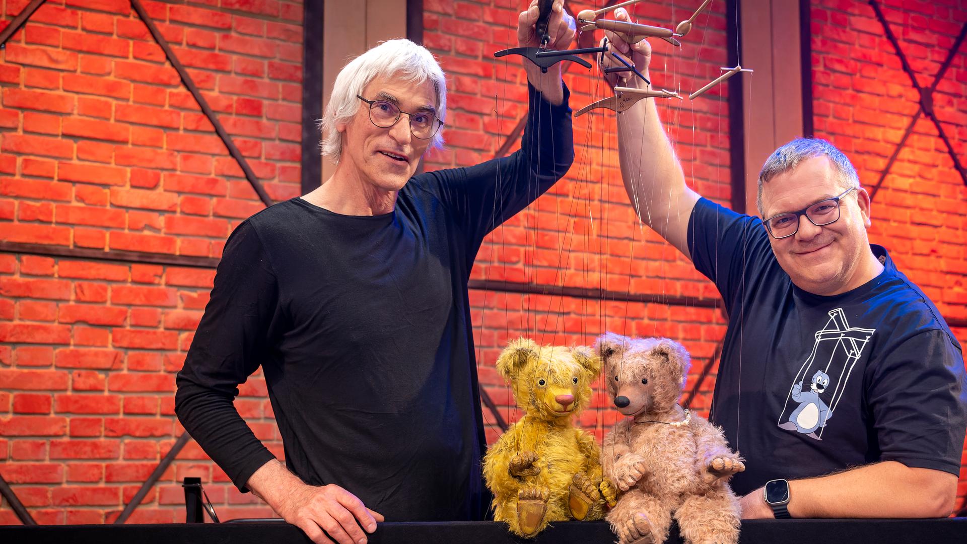 Puppenspieler Detlef Schmelz und Elton stehen nebeneinander und halten Marionetten in den Händen. Die Marionettenpuppen sind zwei Teddybären aus Stoff, die auf einer Kante vor Detlef Schmelz und Elton sitzen.