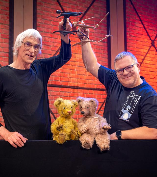 Puppenspieler Detlef Schmelz und Elton stehen nebeneinander und halten Marionetten in den Händen. Die Marionettenpuppen sind zwei Teddybären aus Stoff, die auf einer Kante vor Detlef Schmelz und Elton sitzen.
